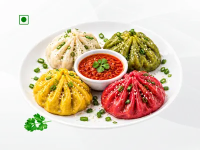 A plate of 4 colorful handcrafted momos in white, green, yellow, and red variants served with spicy chutney.